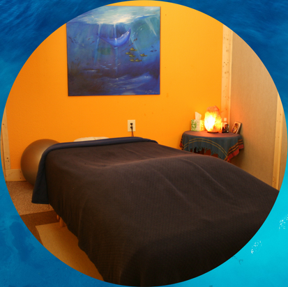 Picture of a comfortable massage space. A soft table with blue flannel sheets, warm orange walls and a lovely painting of the ocean